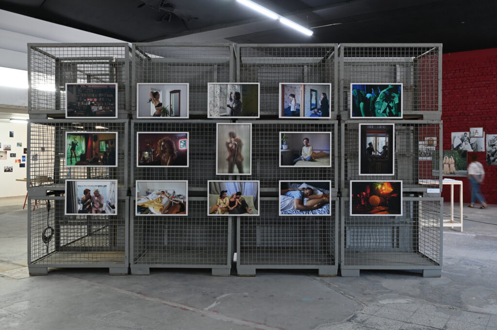 KI generiert: Das Bild zeigt eine Kunstausstellung mit mehreren Fotografien, die an einem Gittergestell angebracht sind. Im Hintergrund sind weitere Kunstwerke und Ausstellungsstücke zu sehen.