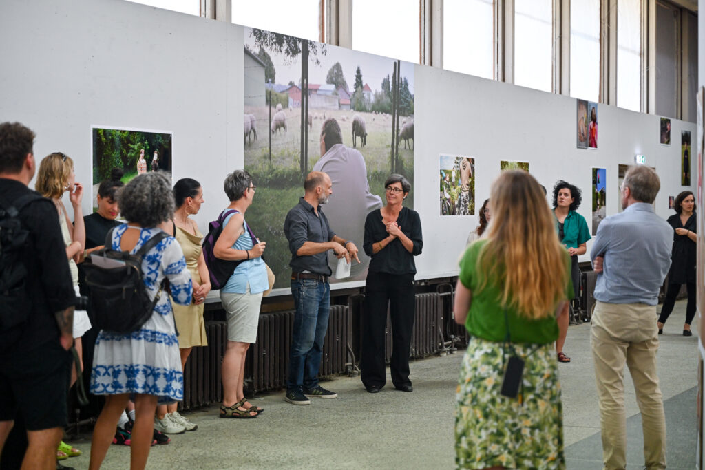 KI generiert: Das Bild zeigt eine Gruppe von Menschen, die sich eine Kunst- oder Fotoausstellung anschauen und miteinander sprechen. Im Hintergrund sind großformatige Fotos an der Wand zu sehen.