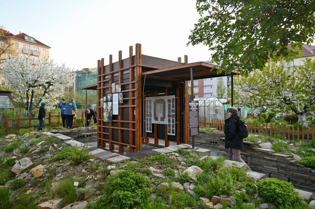 KI generiert: Das Bild zeigt einen kleinen, modernen Pavillon mit Holzstruktur in einem Garten, umgeben von Pflanzen und blühenden Bäumen. Menschen betrachten die ausgestellten Tafeln und erkunden die Umgebung.