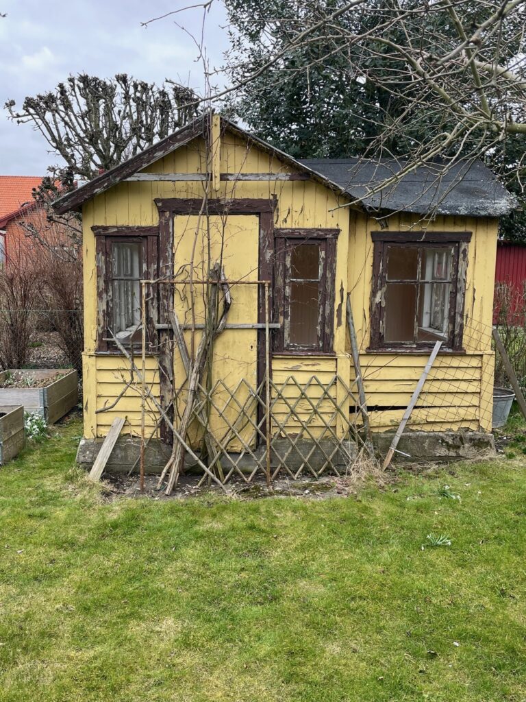 KI generiert: Das Bild zeigt ein altes, gelbes Gartenhaus mit bröckelnder Farbe in einem Garten. Vor dem Haus befinden sich einige Pflanzen und ein Holzspalier.