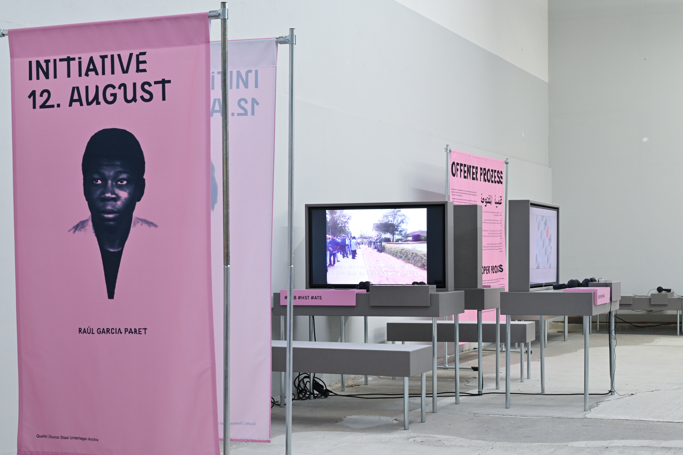 KI generiert: Das Bild zeigt eine Ausstellung, die sich um die "Initiative 12. August" dreht. Verschiedene Informationsstände und ein Bildschirm mit Videoinhalten sind zu sehen, begleitet von Bannern und Plakaten.