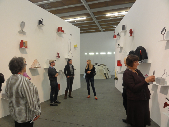 KI generiert: Das Bild zeigt eine Gruppe von Personen, die sich in einer Kunstausstellung befinden. An den Wänden sind verschiedene Kunstwerke und Exponate auf kleinen Regalen ausgestellt.