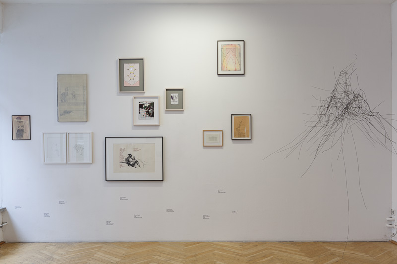 KI generiert: Das Bild zeigt eine Wand in einer Kunstgalerie, an der mehrere gerahmte Kunstwerke in unterschiedlichen Größen und Stilen hängen. Auf der rechten Seite der Wand befindet sich eine drahtartige Skulptur.