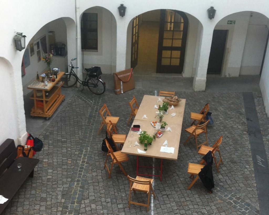 KI generiert: Das Bild zeigt einen Innenhof mit einem großen Tisch in der Mitte, umgeben von mehreren leeren Stühlen. Auf dem Tisch stehen verschiedene Gegenstände wie Pflanzen und Getränke, und im Hintergrund sind ein Fahrrad sowie ein kleiner Servierwagen zu sehen.