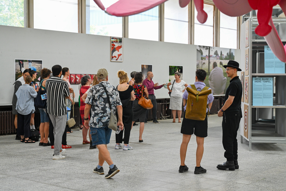 KI generiert: Das Bild zeigt eine Gruppe von Menschen, die sich eine Ausstellung in einer Galerie ansehen. Einige Personen stehen vor Bildern an der Wand, während andere miteinander sprechen.
