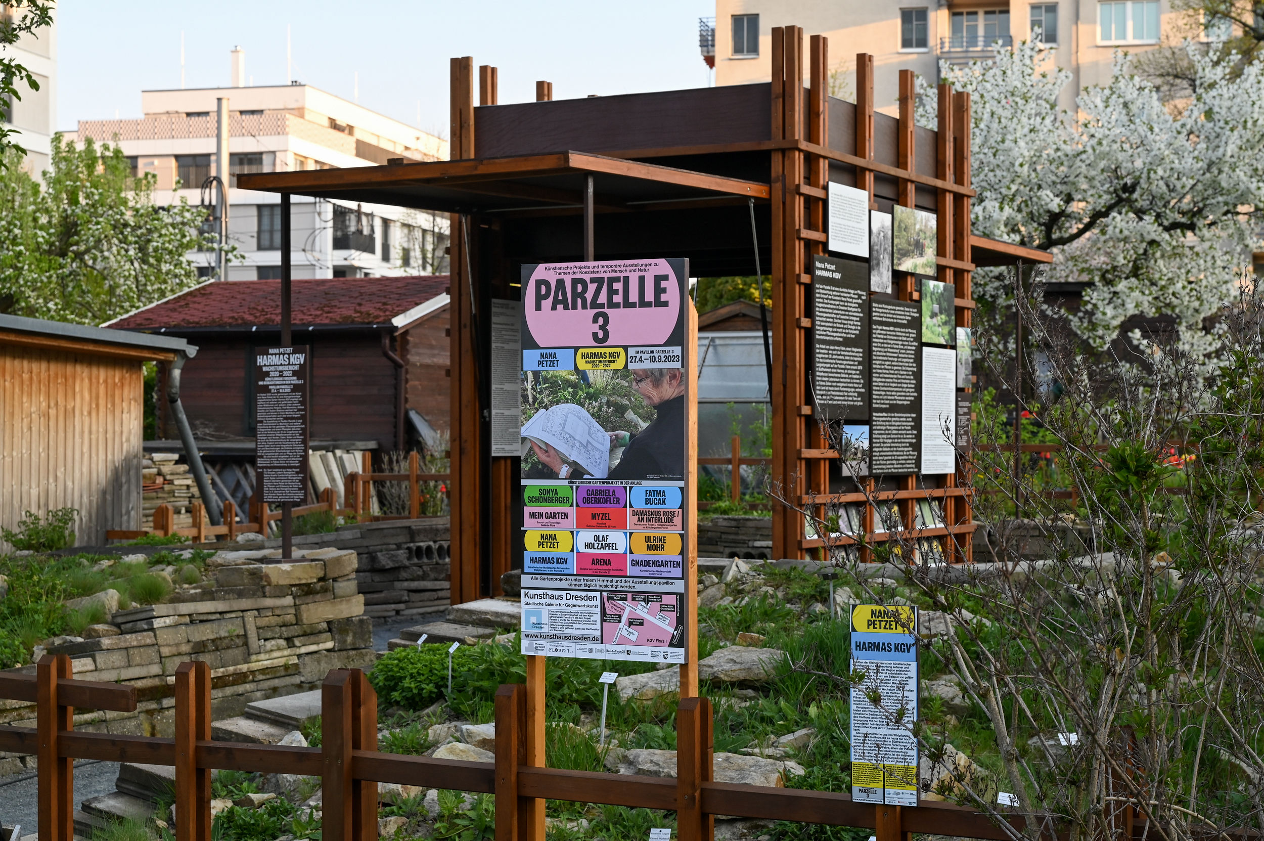 KI generiert: Das Bild zeigt einen Gartenbereich mit mehreren Schildern in deutscher Sprache, darunter ein großes Schild mit der Aufschrift "PARZELLE 3". Im Hintergrund sind Holzkonstruktionen und Informationstafeln zu sehen.