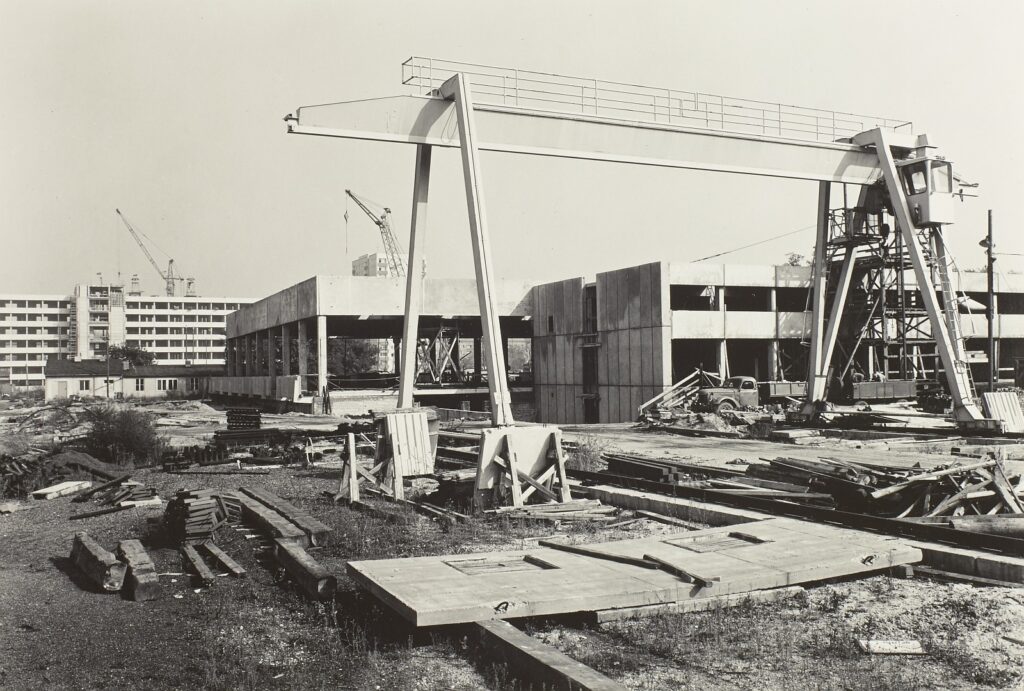 KI generiert: Das Bild zeigt eine Baustelle mit unfertigen Betongebäuden und Baumaterialien im Vordergrund sowie Baukränen im Hintergrund. Der Hauptinhalt ist der Konstruktionsvorgang eines großen Bauprojekts.