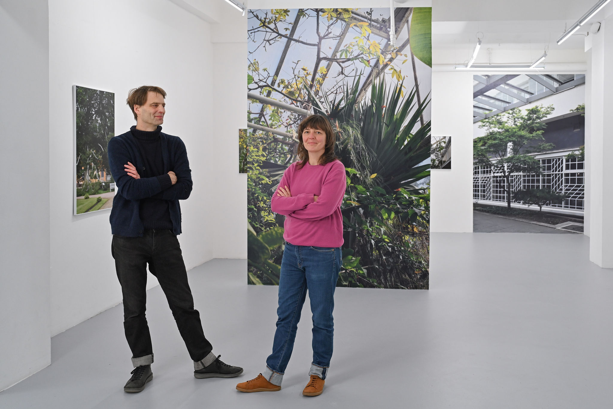 KI generiert: Das Bild zeigt zwei Personen, die in einer Kunstausstellung stehen, umgeben von großformatigen Fotografien, die vorwiegend Pflanzen und Architektur zeigen. Sie stehen entspannt und blicken in Richtung der Kamera.