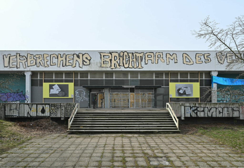 KI generiert: Das Bild zeigt die Außenansicht eines verlassenen Gebäudes, das mit Graffiti bedeckt ist und eine Treppe zum Eingang hat. Auf der Fassade ist groß der Satz "VERBRECHENS BRICHTARM DES..." gesprüht.