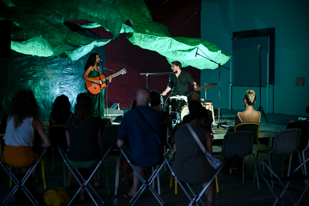 KI generiert: Das Bild zeigt eine Live-Musikdarbietung in einem kleinen, intimen Rahmen, bei der eine Frau Gitarre spielt und singt, während ein Mann Schlagzeug spielt. Einige Zuhörer sitzen auf Stühlen vor der Bühne und lauschen der Aufführung.