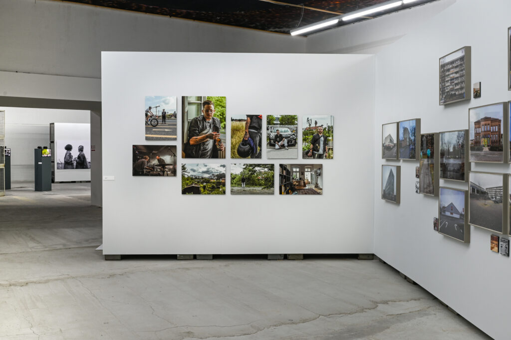 KI generiert: Das Bild zeigt eine Galerieausstellung mit mehreren Fotografien, die an weißen Wänden hängen. Die gezeigten Fotografien umfassen verschiedene Alltagsszenen und Landschaftsaufnahmen.