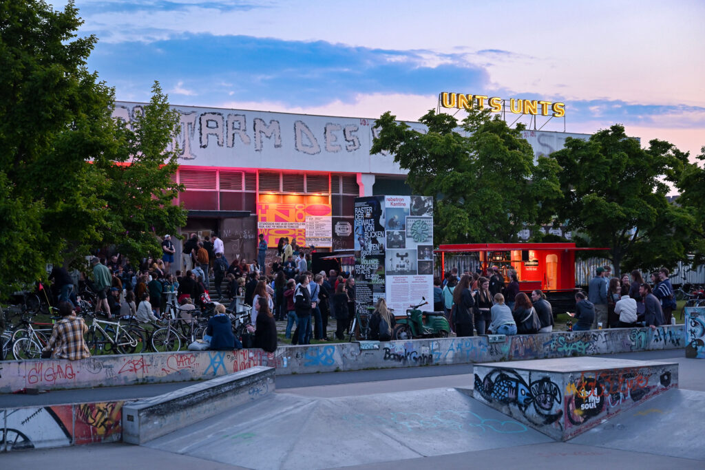 KI generiert: Das Bild zeigt ein urbanes Kulturzentrum oder Event mit vielen Menschen, die sich vor einem Gebäude versammeln, das mit Graffiti und Plakaten bedeckt ist. Im Vordergrund ist ein Skatepark zu sehen, umgeben von Graffiti-bemalten Flächen.