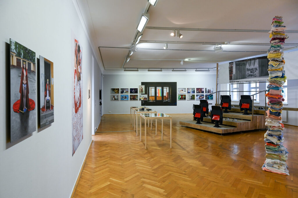 KI generiert: Das Bild zeigt eine moderne Kunstausstellung in einer Galerie, mit Fotografien und Installationen an den Wänden sowie ein Stapel bunter Textilien auf der rechten Seite. In der Mitte des Raumes befinden sich Vitrinentische und eine Sitzgelegenheit mit roten Stühlen.