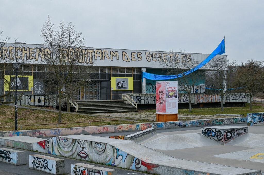 KI generiert: Das Bild zeigt ein graffitiübersätes Gebäude mit dem Schriftzug "ARCHITEKTUR MASSGEBEND IM GEISTE" und einem angrenzenden Skatepark. Im Vordergrund sind farbenfrohe Graffiti und Skaterrampen zu sehen.