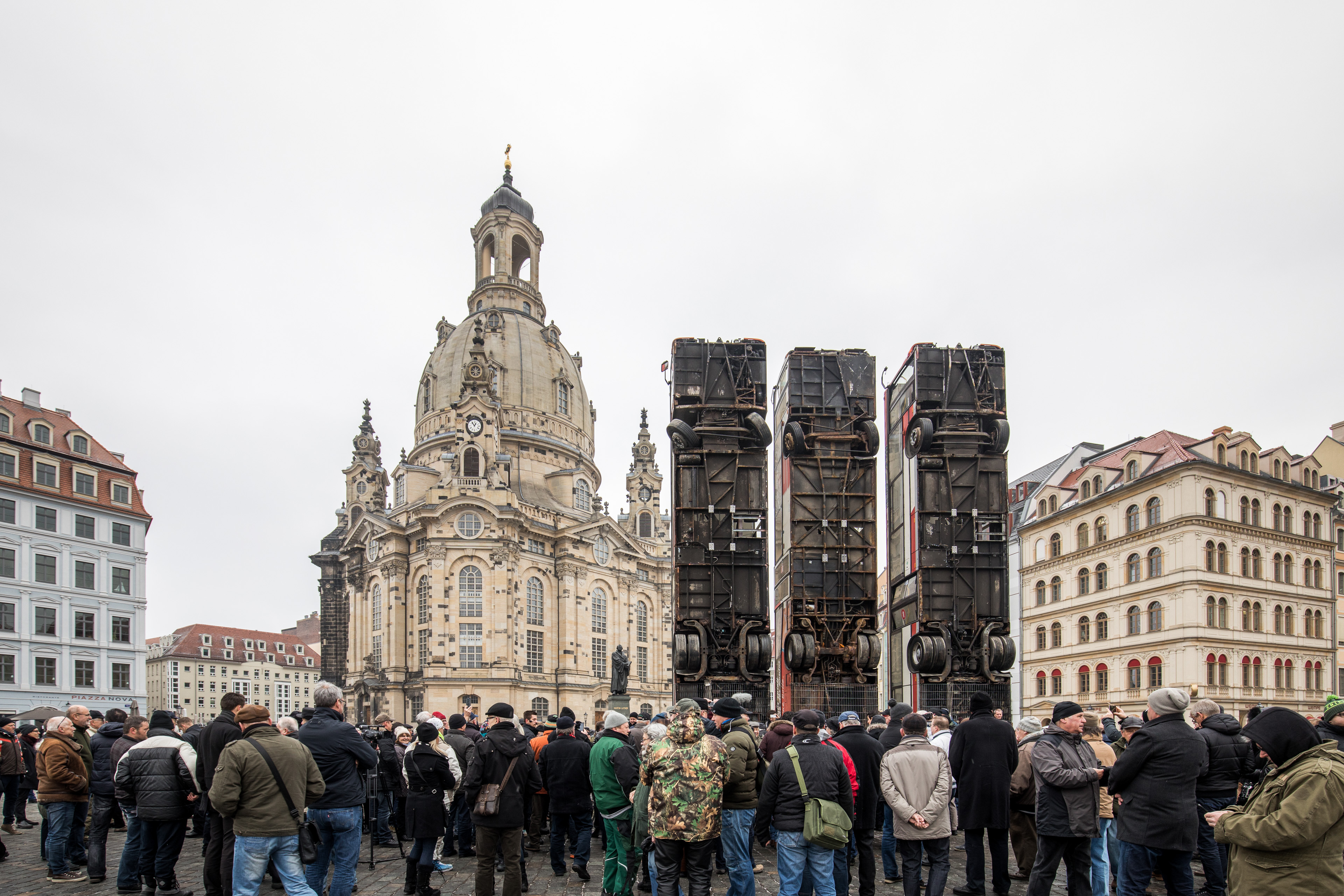 KI generiert: Das Bild zeigt eine Menschenmenge, die sich vor einem historischen Gebäude und drei senkrecht aufgestellten Buswracks versammelt hat. Im Hintergrund ist ein eindrucksvolles Bauwerk mit Kuppel zu sehen, vermutlich eine Kirche.