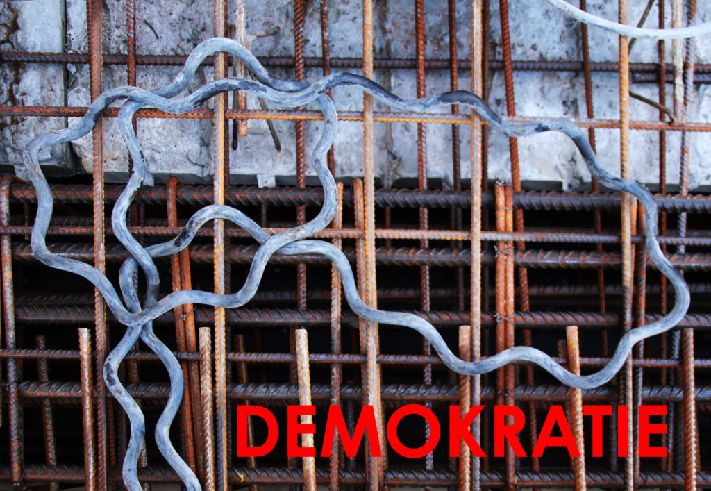 KI generiert: Das Bild zeigt ein unregelmäßiges Metallgebilde vor einem Hintergrund aus rostigen Metallstangen mit dem Wort "DEMOCRATIE" in roten Buchstaben darunter. Es symbolisiert möglicherweise das Thema Demokratie und Struktur.