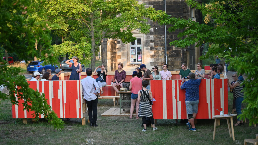 KI generiert: Das Bild zeigt eine Gruppe von Menschen, die sich draußen versammelt haben und miteinander interagieren. Sie stehen und sitzen um eine rot-weiße Struktur in einem grünen, parkähnlichen Bereich.