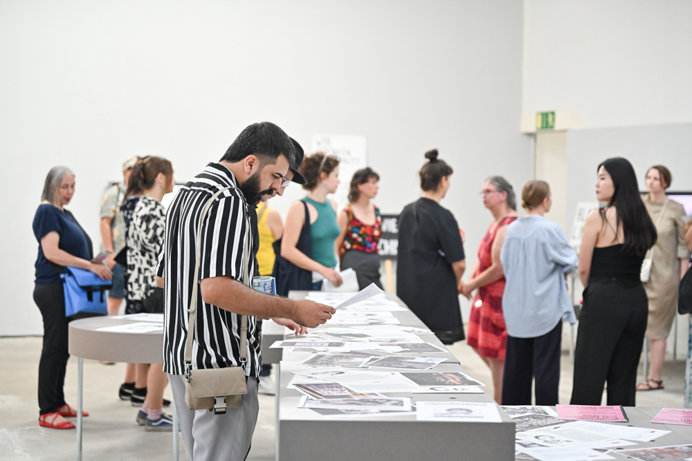 KI generiert: Das Bild zeigt eine Gruppe von Menschen, die sich ein Ausstellung oder eine Präsentation ansehen. Im Vordergrund betrachtet ein Mann aufmerksam die auf einem Tisch ausgestellten Dokumente oder Kunstwerke.