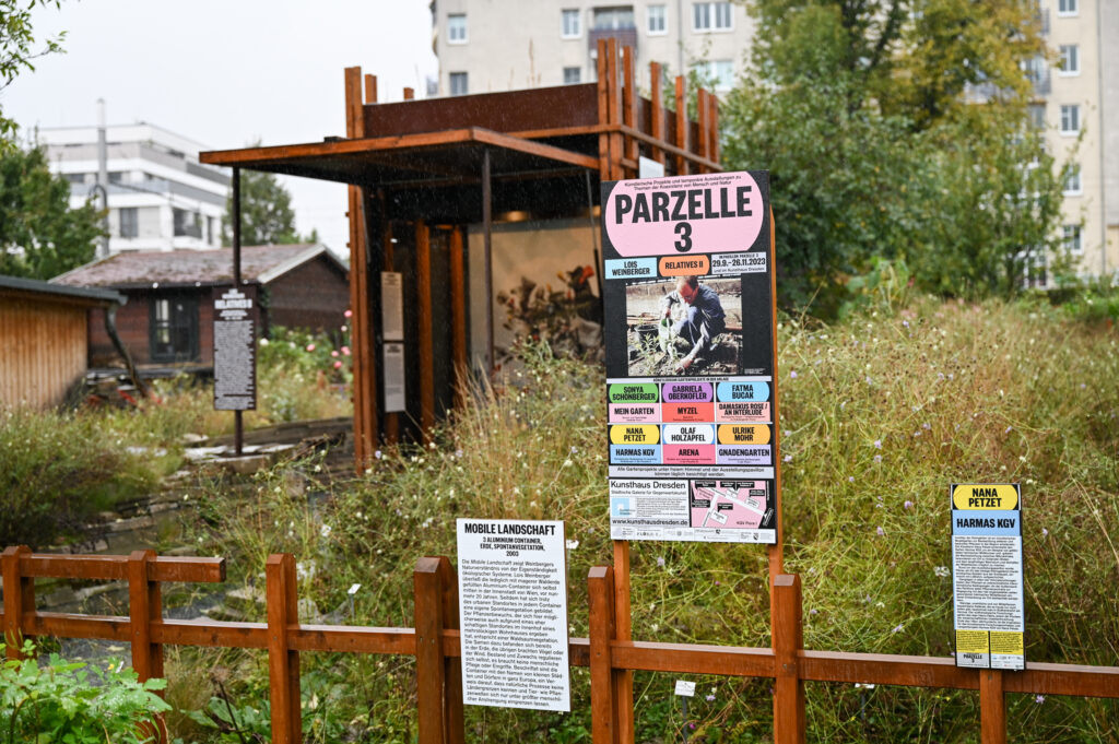 KI generiert: Das Bild zeigt ein urbanes Gartenprojekt mit dem Namen "Parzelle 3", das durch verschiedene Schilder und Beschreibungen vor einem Holzgebäude dargestellt wird. Im Vordergrund steht eine Informationstafel mit detaillierter Beschreibung des Projekts.