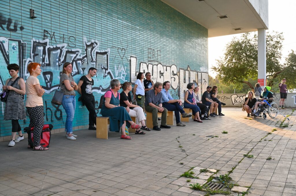 KI generiert: Das Bild zeigt eine Gruppe von Menschen, die in einer Reihe an einer mit Graffiti bedeckten Wand sitzen und stehen. Sie scheinen etwas zu beobachten oder an einer Veranstaltung teilzunehmen.