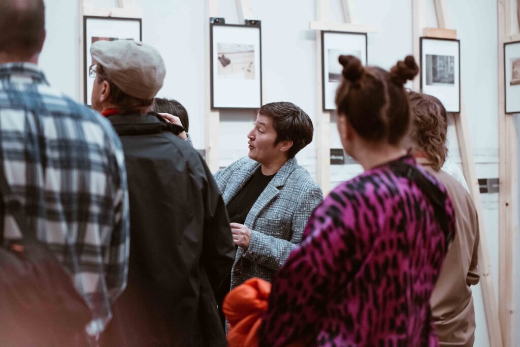 KI generiert: Das Bild zeigt eine Gruppe von Menschen, die Kunstwerke in einer Galerie betrachten. Eine Person in der Mitte scheint zu sprechen und erklärt möglicherweise etwas über die ausgestellten Werke.