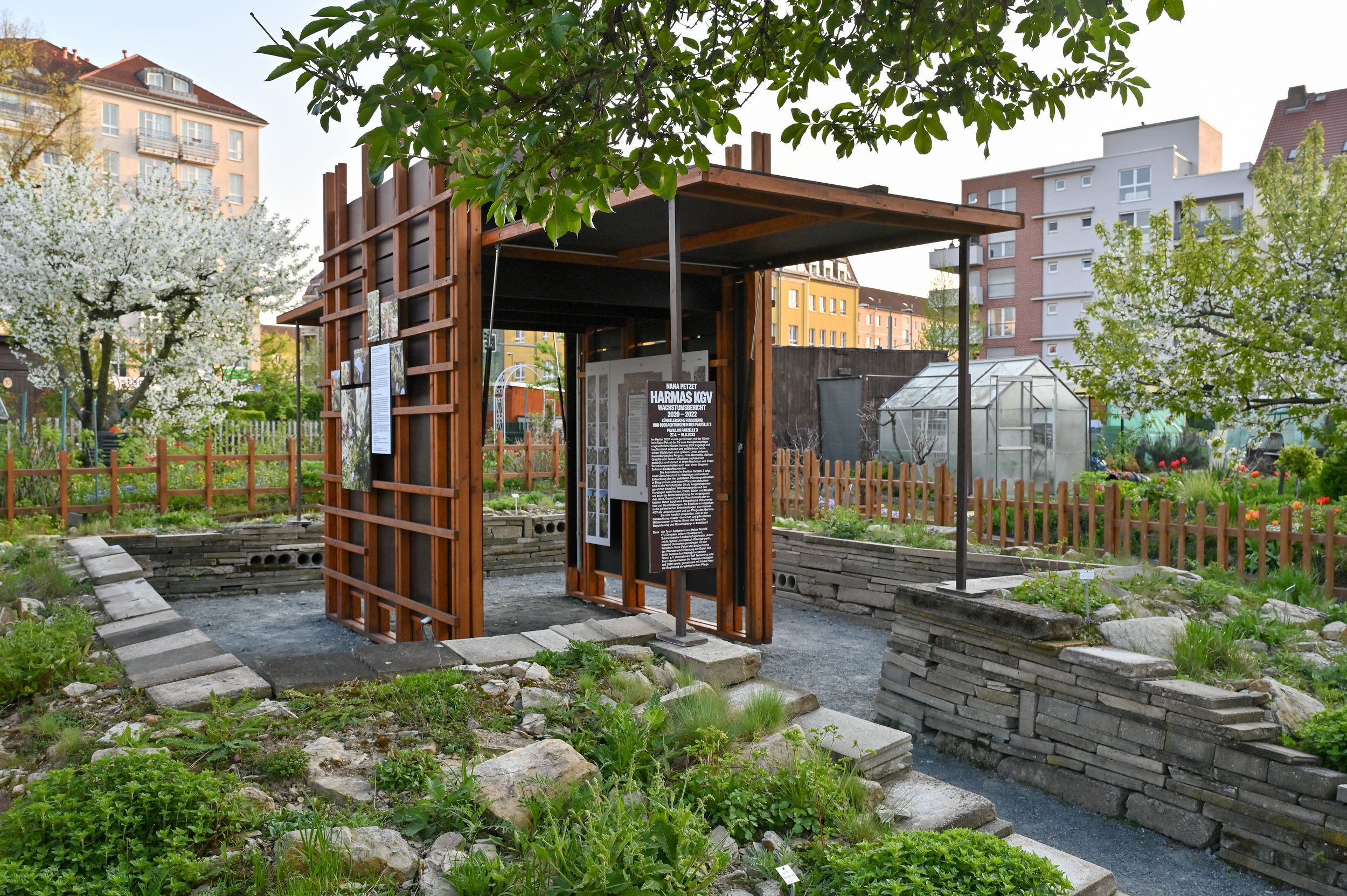 KI generiert: Das Bild zeigt einen kleinen modernen Pavillon in einem städtischen Garten mit informativen Tafeln und umgeben von bepflanzten Beeten und blühenden Bäumen. Im Hintergrund sind Wohngebäude und ein Gewächshaus zu sehen.