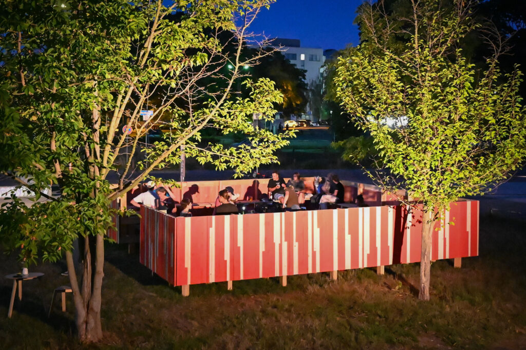 KI generiert: Das Bild zeigt eine Gruppe von Menschen, die abends in einer bunt bemalten Holzkonstruktion im Freien zusammensitzen, umgeben von Bäumen. Es scheint eine gesellige Zusammenkunft oder Veranstaltung zu sein.