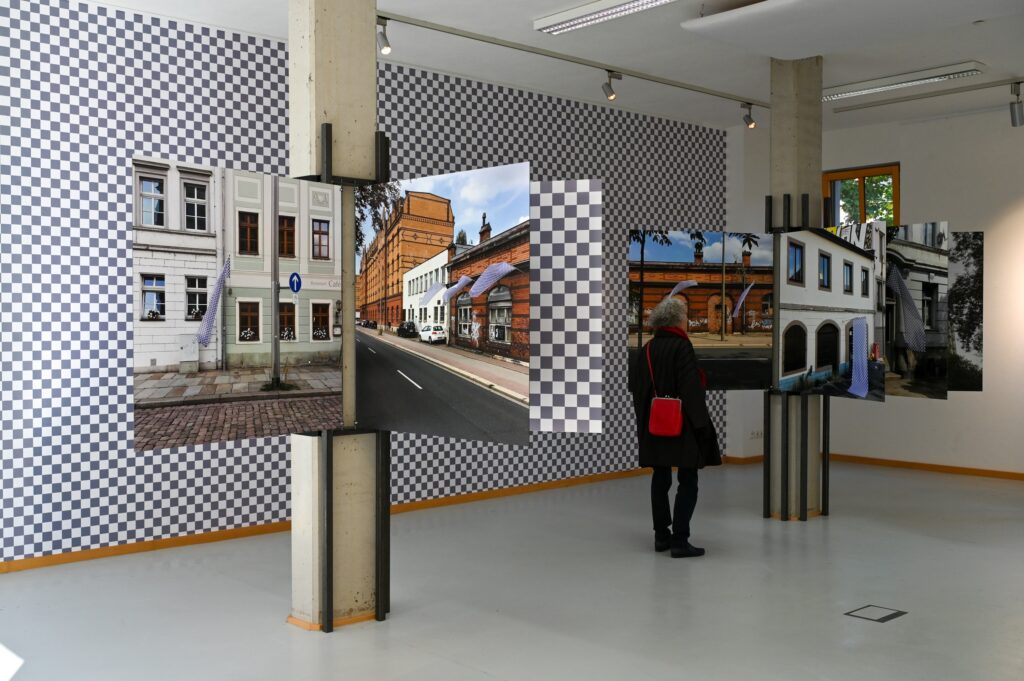 KI generiert: Das Bild zeigt eine Ausstellung in einem modernen, hellen Raum mit einer Person, die mehrere großformatige Fotografien von städtischen Gebäuden betrachtet. Der Hintergrund ist mit einem schwarz-weißen Schachbrettmuster dekoriert.