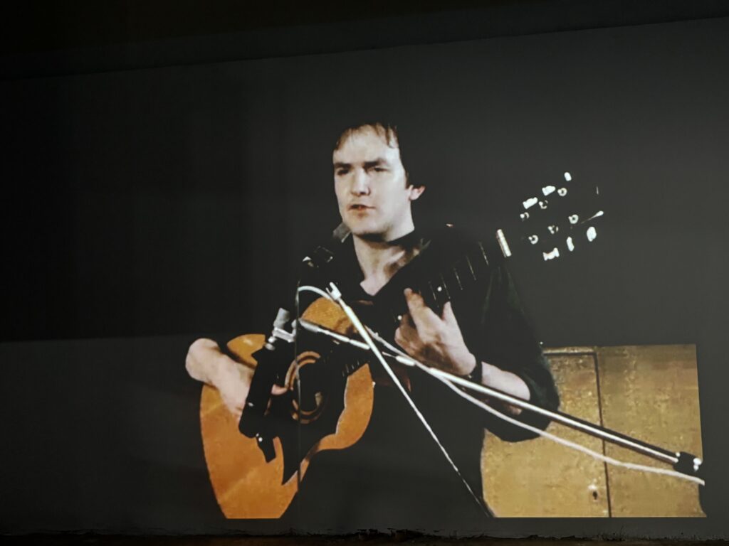 KI generiert: Das Bild zeigt eine Person, die Gitarre spielt und in ein Mikrofon singt. Der Fokus liegt auf der musikalischen Darbietung.