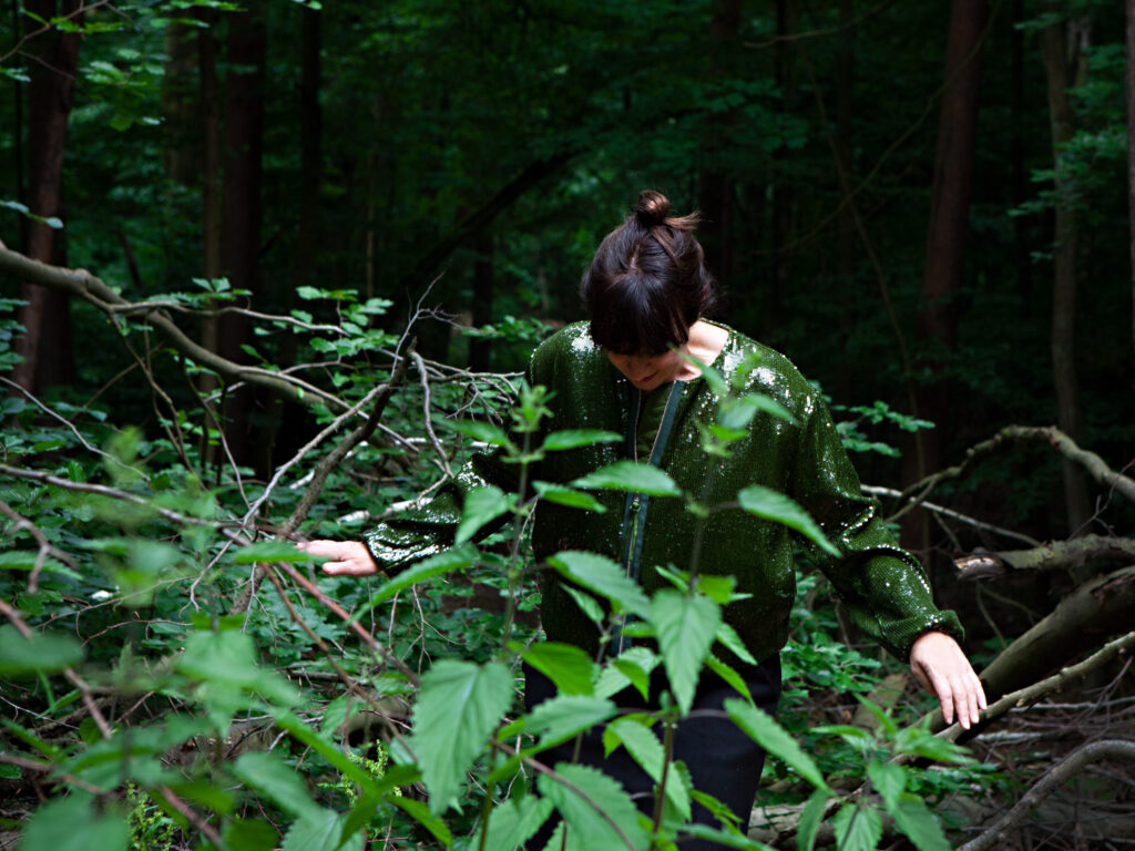 KI generiert: Das Bild zeigt eine Person in einem dichten Wald, die nach unten schaut. Die Person trägt eine glänzende Jacke, die im Kontrast zur natürlichen Umgebung steht.