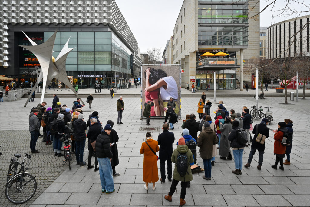 KI generiert: Auf dem Bild versammeln sich Menschen auf einem öffentlichen Platz vor einem großen Foto eines Paares, das sich umarmt. Im Hintergrund sind moderne Gebäude und Geschäfte zu sehen.