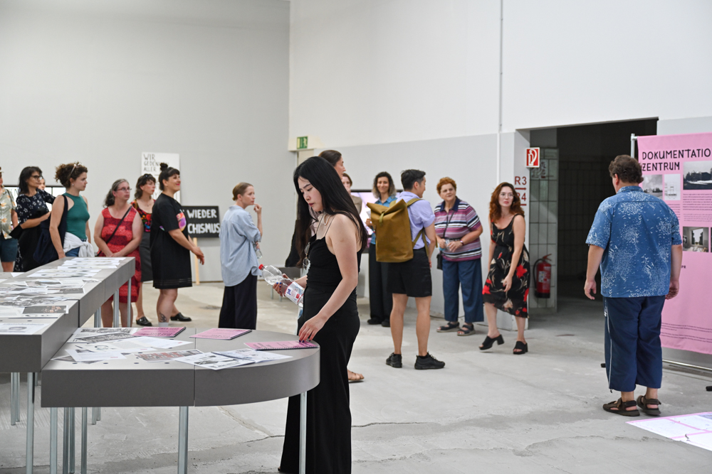 KI generiert: Das Bild zeigt eine Gruppe von Menschen in einer Ausstellungshalle, die verschiedene ausgestellte Dokumente und Plakate betrachten. Im Vordergrund schaut sich eine Frau die ausgestellten Inhalte genauer an.