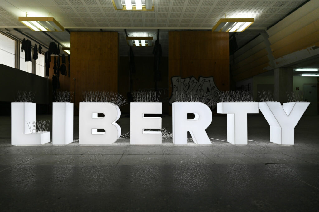 KI generiert: Das Bild zeigt große, weiße Buchstaben, die das Wort "LIBERTY" buchstabieren, in einem Innenraum mit grauem Boden und Deckenbeleuchtung. Im Hintergrund sind Holzwände und Graffitis zu sehen.
