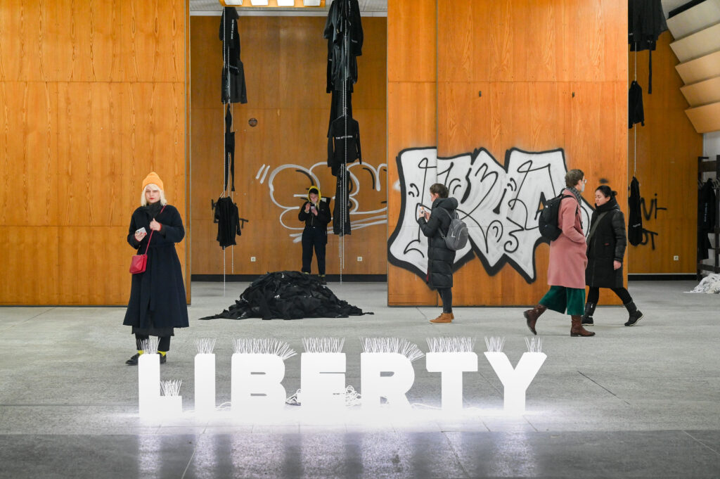 KI generiert: Das Bild zeigt eine Indoor-Ausstellung mit einer leuchtenden Aufschrift "LIBERTY", im Hintergrund sind hölzerne Wände, Graffiti und Kunstinstallationen zu sehen. Mehrere Personen beobachten die Exponate oder gehen durch die Ausstellung.