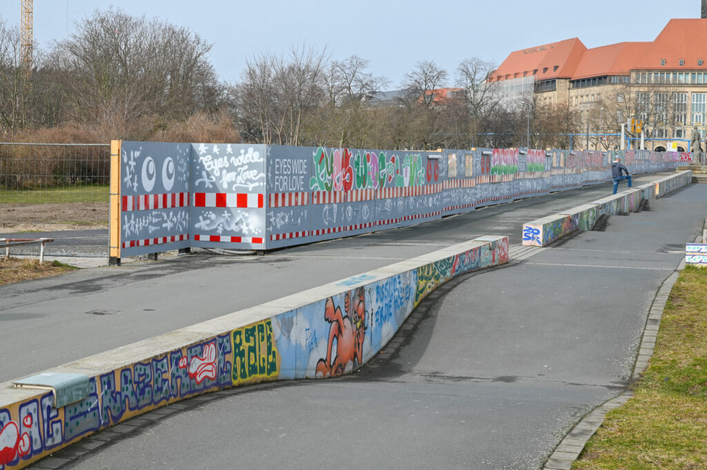 KI generiert: Das Bild zeigt eine mit Graffiti verzierte Betonfläche und eine lange, bemalte Bauzaunbarriere mit dem Schriftzug "Eyes Wide for Love." Im Hintergrund sind Bäume und ein großes Gebäude mit rotem Dach zu sehen.