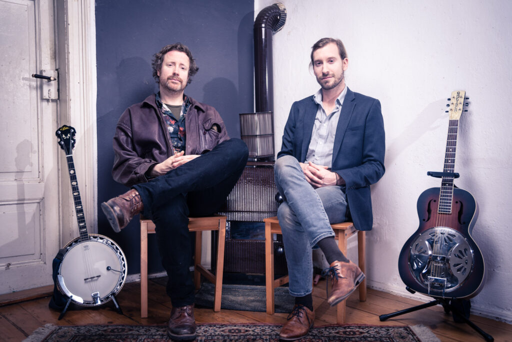 KI generiert: Zwei Männer sitzen in einem Raum mit einem Banjo und einer Resonatorgitarre neben sich. Sie vermitteln eine entspannte, musikalische Atmosphäre.