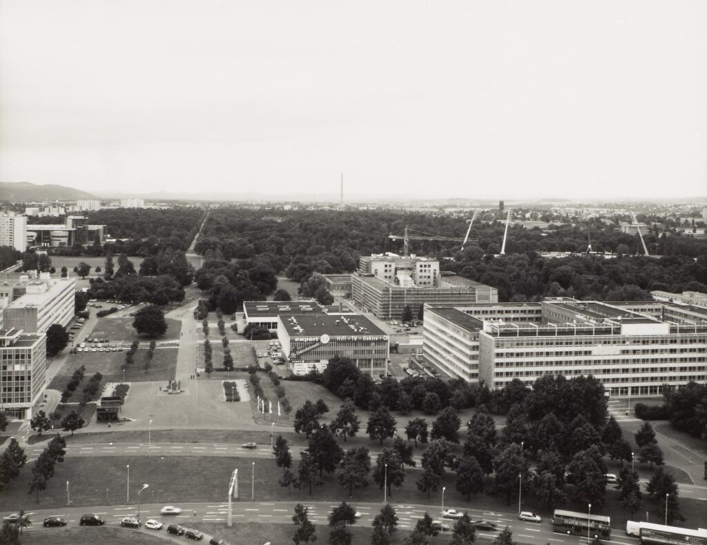 KI generiert: Das Bild zeigt eine städtische Landschaft mit modernen Gebäudekomplexen und einer zentralen Straße, die von Bäumen gesäumt ist. Im Hintergrund erstreckt sich ein großer Park oder Waldgebiet, das die städtische Umgebung ergänzt.