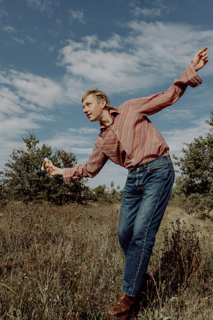 KI generiert: Ein Mann in einem gestreiften Hemd und Jeans posiert dynamisch auf einer Wiesenfläche unter einem teils bewölkten Himmel. In seiner Hand hält er eine kleine Blume, während er sich nach vorn beugt.