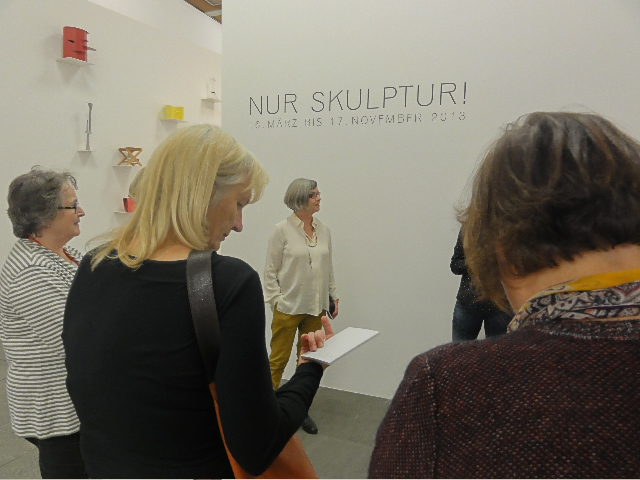 KI generiert: Das Bild zeigt eine Gruppe von Menschen, die eine Ausstellung mit dem Titel "NUR SKULPTUR!" besuchen. Einige der Besucher schauen auf die ausgestellten Werke oder machen sich Notizen.